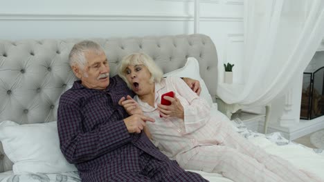 Senior-elderly-couple-wearing-pyjamas-lying-on-bed-looking-on-mobile-phone-making-online-shopping