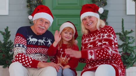 Senior-old-couple-grandparents-with-grandchild-girl-kid-waves-hand-hello,-hi-near-Christmas-house