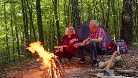 Senior-elderly-grandmother-grandfather-cooking-frying-sausages-over-campfire-in-wood-at-camping