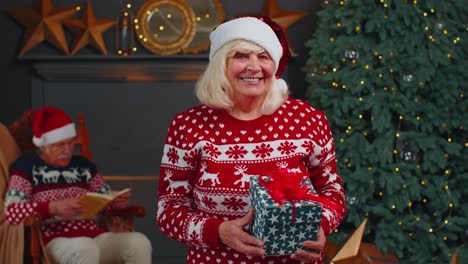 Senior-grandmother-in-festive-sweater-presenting-Christmas-gift-box,-smiling,-looking-at-camera