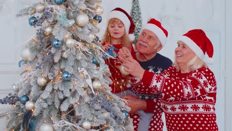 Happy-grandparents,-granddaughter-toddler-kid-hanging-balls-decorating-Christmas-tree-celebrating