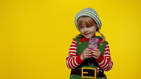 Kid-girl-with-mobile-phone-in-Christmas-elf-Santa-helper-costume-points-fingers-aside-on-blank-wall