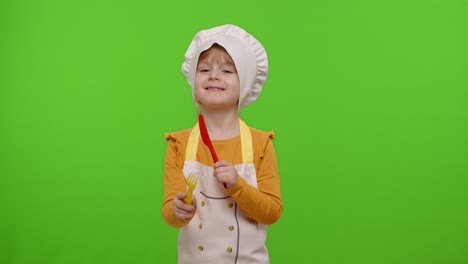 Child-girl-kid-dressed-cook-chef-baker-in-apron-and-hat-with-plastic-fork-and-knife-fooling-around