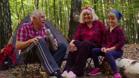 Senior-grandmother-grandfather-granddaughter-telling-funny-stories-fairy-tales-over-campfire-in-wood