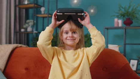 Toddler-girl-sitting-on-home-sofa-using-virtual-reality-headset-helmet-app-to-play-simulation-game