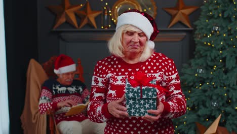 Mature-grandmother-in-festive-sweater-presenting-Christmas-gift-box,-smiling,-looking-at-camera