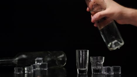 Bartender-pouring-up-two-shots-of-vodka-with-ice-cubes-from-bottle-into-glasses-on-black-background