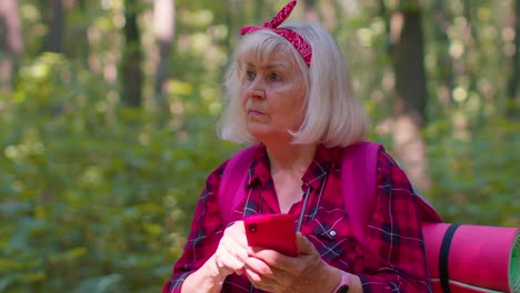 Attractive-senior-old-grandmother-woman-backpacker-planning-trip-route-in-forest-on-mobile-phone