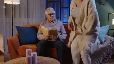 Senior-grandparents-couple-relaxing,-reading-book,-talking-enjoying-leisure-hobbies-at-night-home
