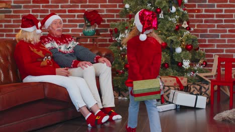 Grandchild-girl-gifting-Christmas-Xmas-present-box-to-amazed-happy-grandparents-at-home,-celebrating