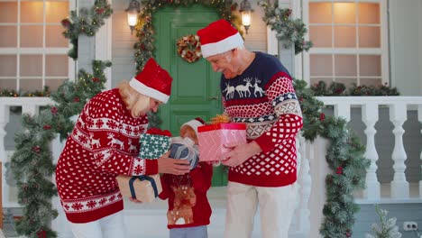 Senior-grandparents-with-grandchild-girl-kid-exhcanging-gifts-near-Christmas-house-celebrating-Xmas