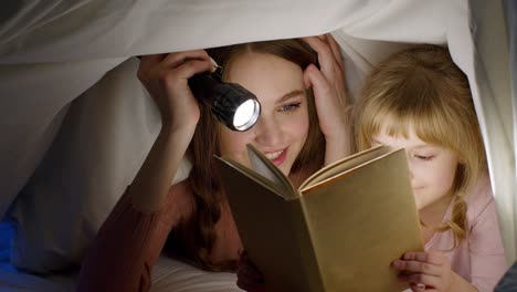 Young-mother-reading-goodnight-story-fairytale-to-child-daughter-under-duvet-blanket-in-night-room