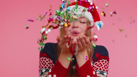 Cheerful-smiling-girl-in-Christmas-Santa-sweater-blowing-on-gold-glitter-confetti-towards-to-camera