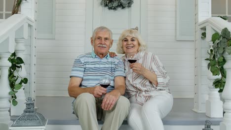 Atractiva-Pareja-De-Ancianos-Caucásicos-Sentados-Y-Bebiendo-Vino-En-El-Porche-De-Casa,-Haciendo-Un-Beso