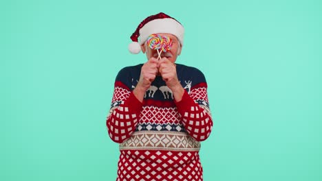 Grandfather-in-Christmas-sweater-holding-candy-striped-lollipops-hiding-behind-them,-fooling-around