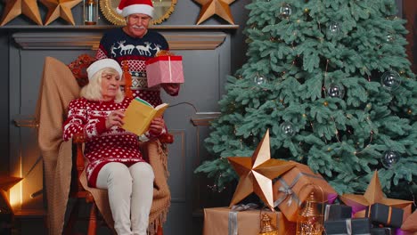 Senior-grandmother-presenting-Christmas-gift-box-to-surprised-grandfather-while-she-reading-book
