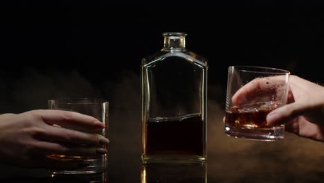 Two-hands-with-glasses-of-cognac-whiskey-with-ice-cubes-making-cheers-toast-on-black-background
