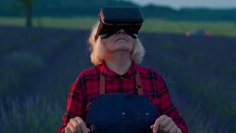 Senior-grandmother-farmer-in-virtual-reality-helmet-managing-quality-in-field-of-lavender-flowers