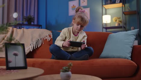 Worried-child-girl-kid-enthusiastically-playing-racing-video-games-on-mobile-phone-at-home-on-sofa
