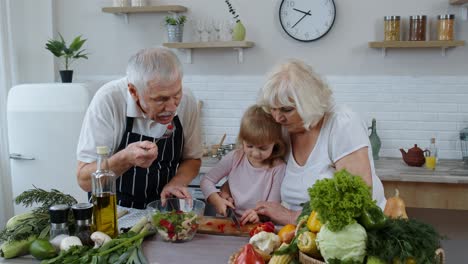Senior-couple-in-kitchen-teaching-granddaughter-child-how-to-cook,-chopping-pepper-with-knife