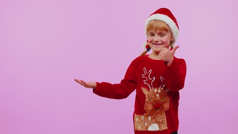 Girl-child-in-Christmas-sweater-deer-showing-thumbs-up-and-pointing-at-on-blank-advertisement-space