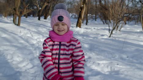 Child-girl-running-on-snowy-road,-fooling-around,-smiling,-looking-at-camera-in-winter-park-forest
