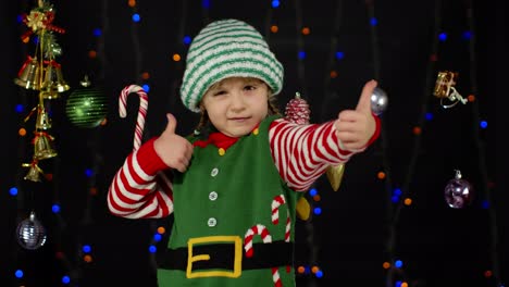Teen-kid-girl-in-Christmas-elf-Santa-Claus-helper-costume-showing-thumbs-up-on-black-background