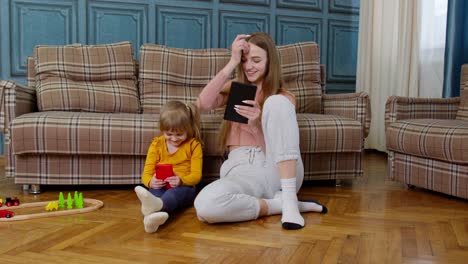 Woman-nanny-and-child-girl-studying-together-with-mobile-phone-and-tablet,-sitting-on-floor-at-home