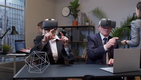 Elderly-old-man-boss-with-woman-colleague-wearing-virtual-reality-glasses-tries-3D-app-for-VR-helmet