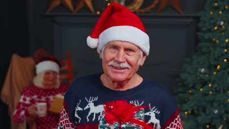 Senior-grandfather-in-festive-sweater-presenting-Christmas-gift-box,-smiling,-looking-at-camera