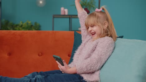 Glückliches-Kleines-Schulkind-Mädchen-Nutzt-Handy-Und-Macht-Gewinnergeste,-Um-Allein-Auf-Dem-Sofa-Zu-Hause-Zu-Feiern
