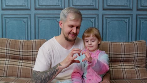 Father-and-child-daughter-kid-in-pajamas-sit-on-couch-smiling,-making-sign-of-shape-heart-near-chest
