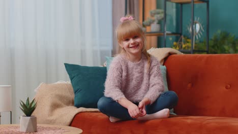 Portrait-of-little-adorable-child-girl-sitting-alone-on-sofa-at-home-looking-at-camera-and-smiling