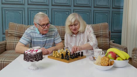 Retired-senior-couple-talking-drinking-tea,-playing-chess-in-modern-living-home-room-lounge-together