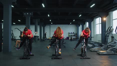 Group-athletic-girls-performing-aerobic-riding-training-exercises-on-cycling-stationary-bike-in-gym