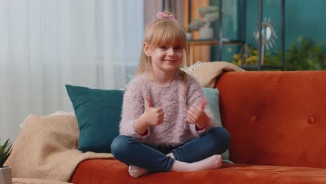 Child-girl-kid-sitting-on-sofa-at-home-alone-showing-thumbs-up-like-sign-positive-something-good
