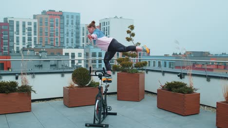 Sportliches-Mädchen,-Das-Aerobic-Trainingsübungen-Auf-Einem-Stationären-Fahrrad-Auf-Dem-Hausdach-Durchführt