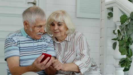 Senior-elderly-caucasian-couple-using-mobile-phone-in-porch-at-home.-Enjoy-online-shopping,-news