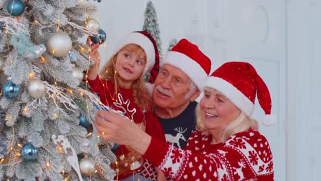 Toddler-girl-with-senior-couple-grandparents-decorating-artificial-Christmas-pine-tree-at-home-room