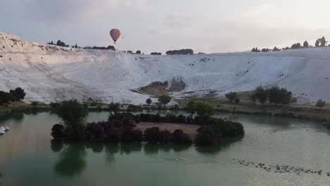 Aerial-view-of-lake-surrounded-by-limestone-formations-with-hot-air-balloon-flying-in-background-in-Pamukkale,-Turkey