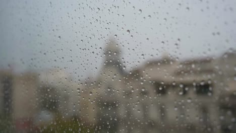 Eine-Schmale-Zeitlupenaufnahme-Von-Regentropfen,-Die-Bei-Starkem-Regen-Durch-Ein-Fenster-Gesehen-Werden,-Mit-Einer-Städtischen-Stadtlandschaft-Im-Hintergrund