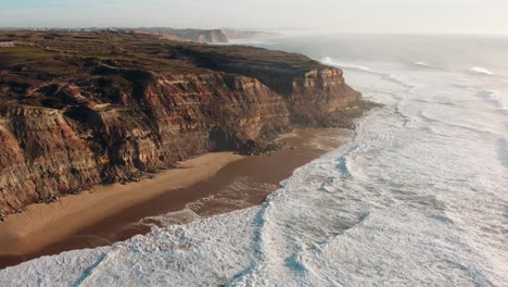Aerial-of-south-imposing-cliffs-rising-straight-up-from-the-beach,-rugged-and-scenic-headland-coast-of-Portugal,-the-westernmost-point-in-continental-Europe