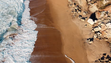 Top-aerial-of-sea-waves-crashing-into-a-majestic-beach-with-mixture-of-sand-and-rocks-on-the-coastline
