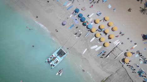 Luftaufnahme-Eines-Weißen-Strandes-Voller-Bunter-Sonnenschirme-Und-Entspannter-Menschen,-Die-Auf-Einem-Klaren-Meer-Schwimmen