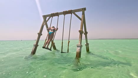 A-woman-in-a-light-blue-bikini-on-a-wooden-swing-in-clear-water,-Egypt