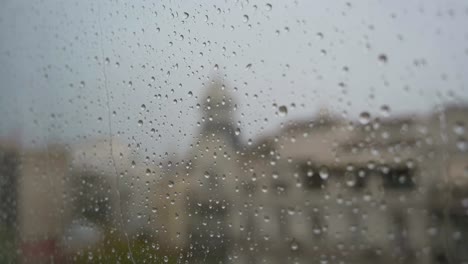 A-slow-motion-footage-of-raindrops-seen-through-a-window-during-heavy-rainfall,-with-an-urban-city-landscape-in-the-background
