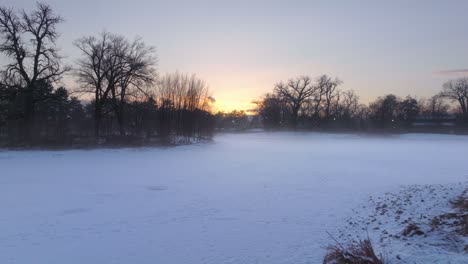Aerial-dolly-over-snow-covered-wooded-banks-and-park-as-sun-rises-shining-light-on-mystic-fog