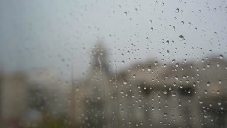 A-slow-motion-of-raindrops-seen-through-a-window-during-heavy-rainfall,-with-an-urban-city-landscape-in-the-background
