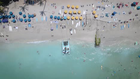 Aerial-footage-captures-the-beauty-of-a-white-sandy-beach-filled-with-lively-beach-umbrellas,-and-people-indulging-in-a-refreshing-swim