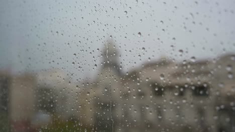 A-slow-motion-view-of-raindrops-seen-through-a-window-during-heavy-rainfall,-with-an-urban-city-landscape-in-the-background